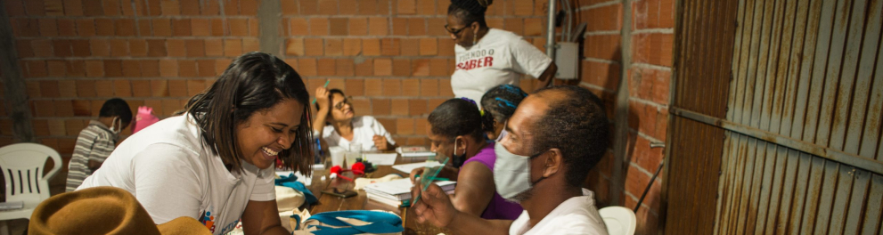 Mulher sorri diante de um senhor que está sentado do outro lado de uma bancada em que outros adultos estão estudando.