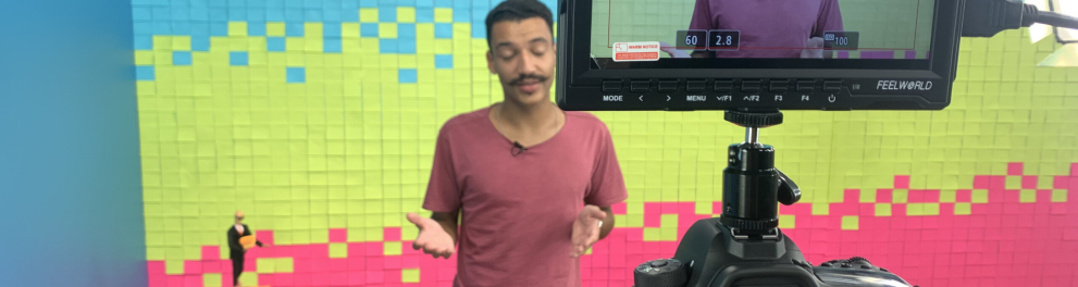 Homem magro e branco, de bigode, está diante de uma câmera de filmar falando. É o apresentador de um programa de TV e está no estúdio. Atrás dele podemos notar o cenário colorido em azul claro, rosa e um tom de amarelo
