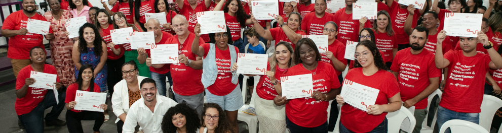 Diversas pessoas de camisas vermelhas seguram seus certificados de conclusão de ensino em cerimônia de formatura.