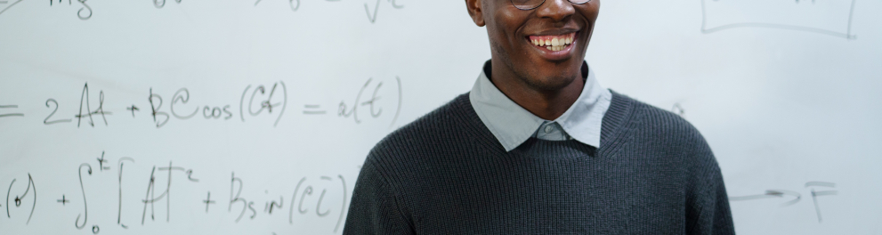 Imagem mostra um Homem sorrindo, de óculos, em frente a quadro branco com equações escritas nele
