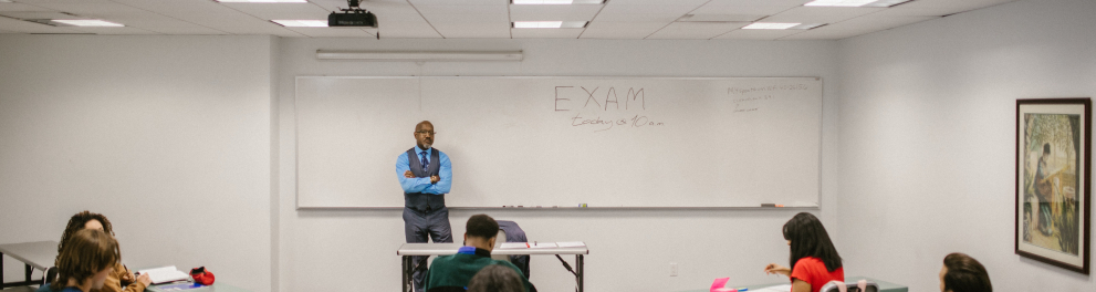 Imagem mostra um professor em frente a um quadro branco, diante dos alunos, numa sala de aula.