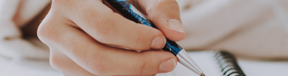 Imagem mostra uma mão segurando uma caneta sobre um caderno.