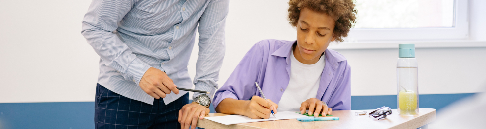Imagem mostra um homem orientando garoto enquanto ele escreve em caderno.