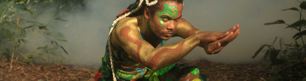 Um homem negro com tranças compridas está ajoelhado e com o corpo todo coberto por folhas verdes e tintas coloridas, onde predominas as cores verde e amarela. Ele está com os braços esticados à frente e com a palma da mão direita em cima do dorso da mão esquerda