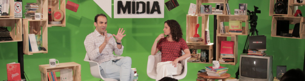 Imagem do programa de TV Idade Mídia. Há um homem branco sentado ao lado de uma mulher branca sentada e, ao fundo, está o cenário com o logotipo do programa em uma parede verde e várias estantes com livros e outros objetos de comunicação, como uma câmera de TV, uma máquina de datilografia, etc
