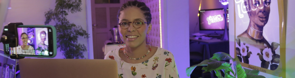 Imagem de uma mulher negra, de cabelos bem curtos e óculos de grau. Ela está sorridente no centro da imagem, com um notebook aberto à sua frente e o cenário do programa Cineclube Futura ao fundo