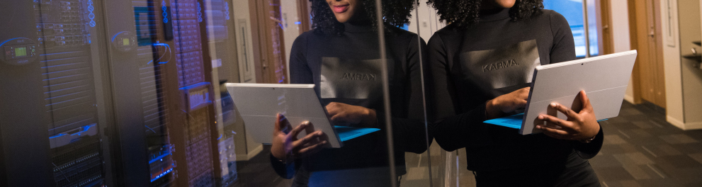 Uma linda mulher negra de cabelo encaracolado vestindo calça e blusa pretas está em pé segurando um notebook com o braço esquerdo e teclando nele com a mão direita, enquanto está encostada em uma vidraça refletindo sua imagem