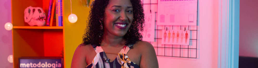 Mulher negra sorridente, de cabelos cacheados está olhando para quem bateu a foto. Ela está no estúdio do programa E aí, professor? e, ao fundo, podemos ver uma estante amarela com acessórios de educação, além de um painel com a palavra "metodologia"