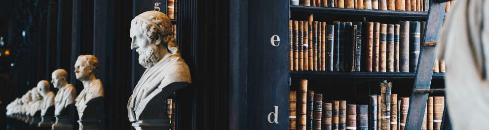 Imagem de uma biblioteca, pois aparecem vários livros em estantes enormes e bustos de filósofos famosos historicamente estão enfileirados ao lado esquerdo