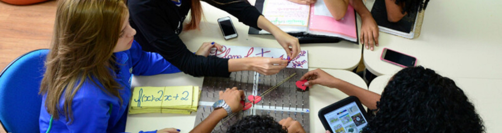 Seis alunos uniformizados estão sentados ao redor de uma mesa e mexendo em tablets, cadernos e um trabalho artesanal em cima da mesa