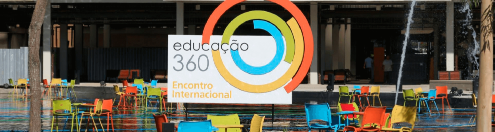 Imagem de um espaço ao ar livre, com gramado verde e várias cadeiras coloridas e mesinhas. No centro da imagem há uma espécie de outdoor com a logomarca do Educação 360