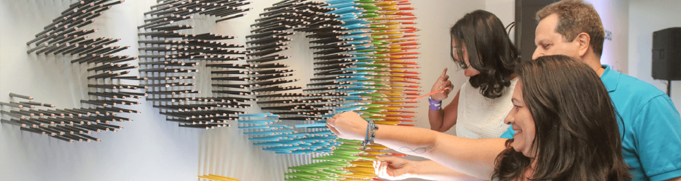 Um homem e duas mulheres estão inserindo lápis coloridos em um mural que está formando a logomarca da Educação 360