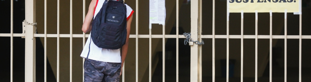 Um estudante está uniformizado e de mochila às costas parado em frente à entrada do colégio, que está com as portas fechadas e um cartaz escrito "Aulas suspensas"