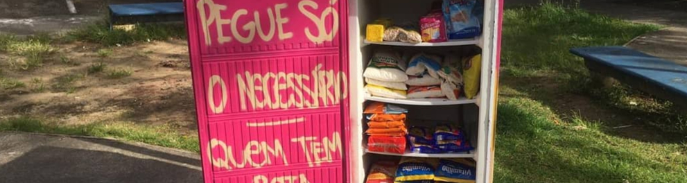 Uma geladeira com a porta cor de rosa e os dizeres "Peque só o necessário / Quem tem, bota. Quem não tem, pega". A geladeira está ao ar livre,, no meio do que parece ser um parque e está com a porta aberta, revelando seu interior repleto de alimentos não perecíveis