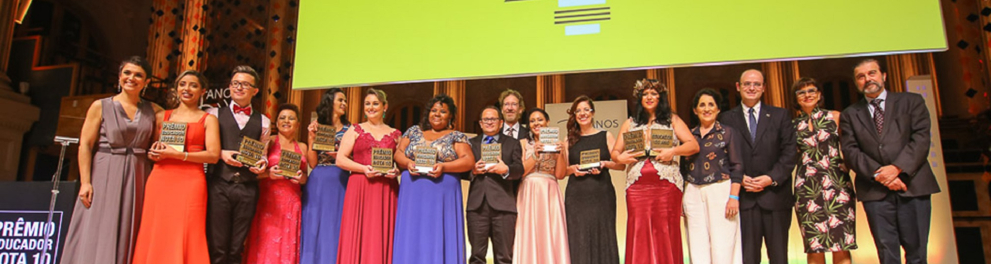 Imagem de várias pessoas lado a lado em cima de um palco, vestidos de forma bem elegante e com troféus em suas mãos. É uma cerimônia de premiação