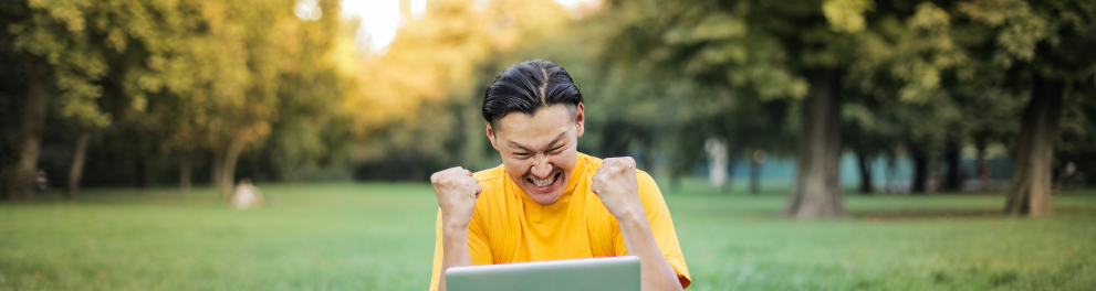 Um adolescente oriental está sentado em um gramado que parece ser um parque, já que há dezenas de árvores ao fundo e nas laterais. Ele está comemorando e olhando feliz para o notebook aberto em seu colo