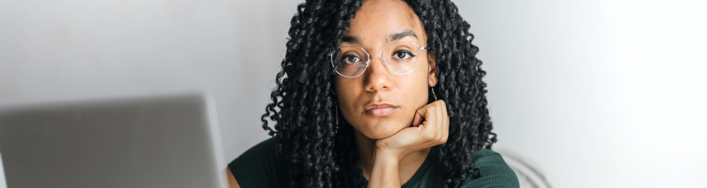 Uma linda e jovem mulher negra de cabelos escuros encaracolados está sentada apoiando o cotovelo esquerdo na mesa e a cabeça em cima da palma da mão esquerda. Ela está em frente a um notebook aberto