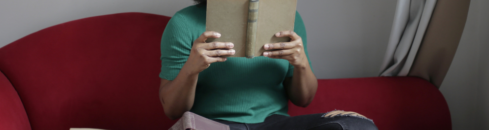 Mulher negra está sentada em um sofá de cor vinho com vários livros ao seu redor e um em sua mãos, aberto e sendo lido