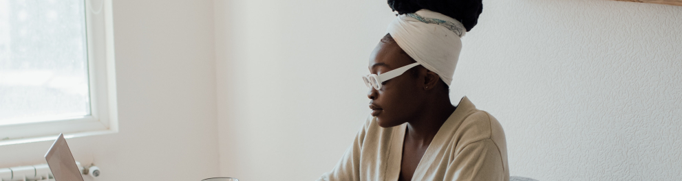 Uma mulher negra sentada em um escritório escrevendo em um caderno e usando um notebook
