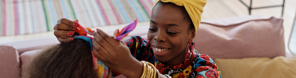 Uma mulher e uma criança negra. A mulher está ajudando a criança a colocar um lenço na cabeça.