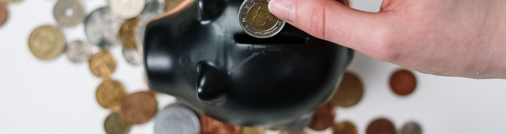 imagem da mão de uma pessoa branca inserindo uma moeda dourada dentro de um cofrinho preto de porquinho. Várias moedas estão espalhadas ao redor do porquinho