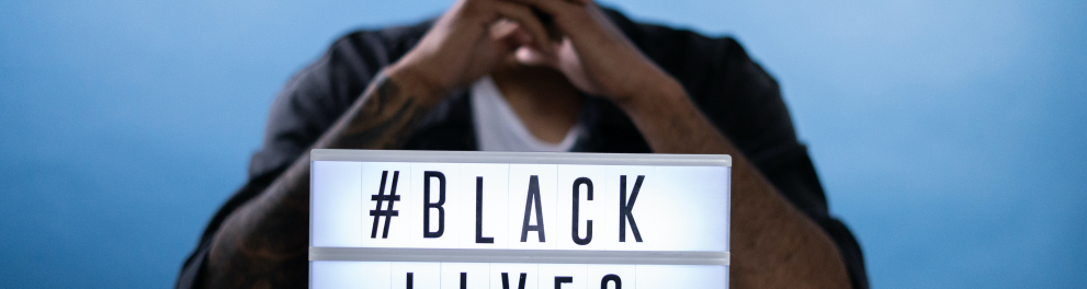 Homem negro sentado e com os cotovelos apoiados em uma mesa. Entre seus cotovelos está um letreiro com os dizeres "#Black Lives Matter"