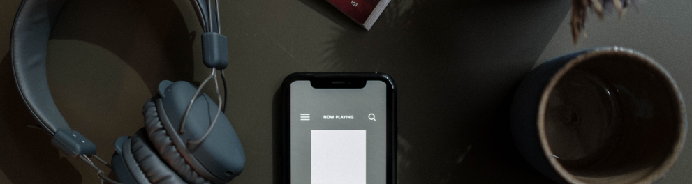 Um celular ligado em uma tela mostrando um podcast, um headphone e uma xícara estão em cima de uma mesa