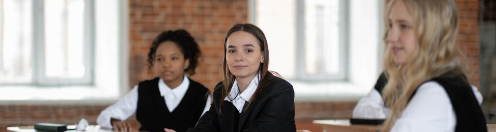 Três meninas uniformizadas estão sentadas em sala de aula, lado a lado. Uma delas está olhando para quem bateu a foto