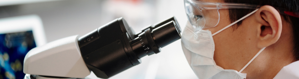 Um cientista de máscara branca e óculos de proteção está usado um microscópio e olhando através de suas lentes