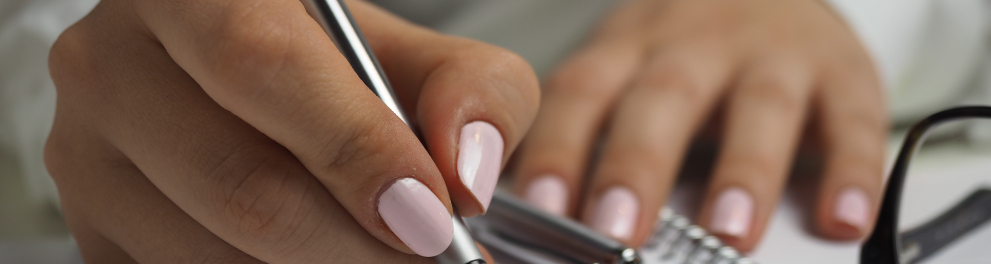 Imagem das mãos de uma mulher branca escrevendo em uma folha de um caderno com uma caneta prateada. As mãos têm unhas compridas e pintadas de rosa claro