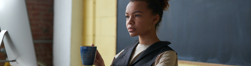 Uma jovem mulher negra está sentada à mesa, que contém dois livros e um computador branco. A moça está com a mão esquerda no teclado do computador e segurando uma caneca com a mão direita. Seu semblante é de seriedade