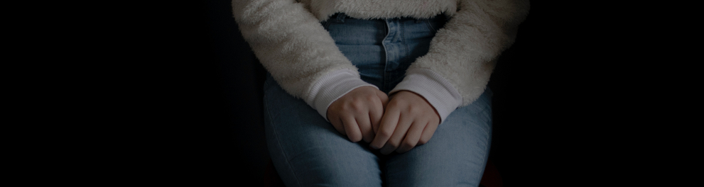 Imagem de uma mulher branca sentada em uma cadeira. A imagem está cortando o rosto da moça, então só vemos do tronco para baixo. Ela está de calça jeans e blusa de manga comprida branca, com as duas mãos fechadas sobre as coxas. O fundo é todo preto