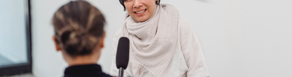 Uma mulher, de costas para quem fotografou, segura o microfone para entrevistar outra mulher, esta de frente para o fotógrafo. A entrevistada está sorrindo.