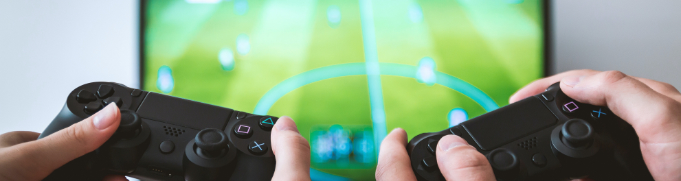 Imagem em close das mãos de duas pessoas segurando controles de video game e jogando um jogo de futebol na TV que está à frente