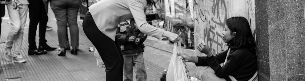 Foto em preto e branco de uma mulher entregando uma refeição embrulhada em um saco plástico a uma moradora de rua, que está sentada em uma calçada e esticando o braço para receber a oferenda