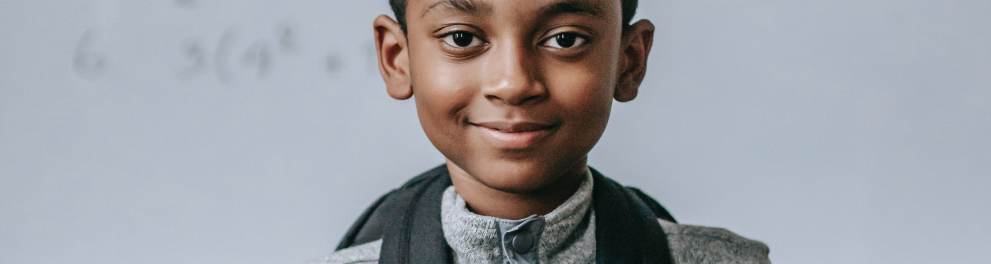 Menino negro sorrindo para quem bateu a foto. Ele está de mochila às costas e segurando as alças com ambas as mãos. Ao fundo, notamos um quadro de sala de aula