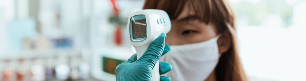 Uma moça oriental está de máscara de prevenção à covid-19 e tendo sua temperatura verificada por um termômetro em formato de pistola direcionado para sua testa