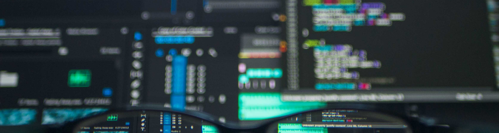 A imagem mostra um óculos de grau com armação preta posicionado em cima de uma mesa. Atrás das lentes do óculos estão algumas telas de computadores, todas conectadas em dados de programação