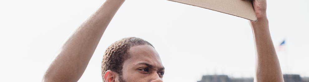 Homem negro, barbudo e de cabelo raspado baixinho está ao ar livre, possivelmente sobre uma ponte, segurando um cartaz com os dizeres "human rights". Ele está sério e trajando camisa cinza