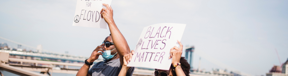 Um homem e uma mulher negra estão usando máscaras de prevenção à covid e segurando cartazes de protesto contra o racismo acima de suas cabeças. Eles estão ao ar livre e parece em cima de uma ponte