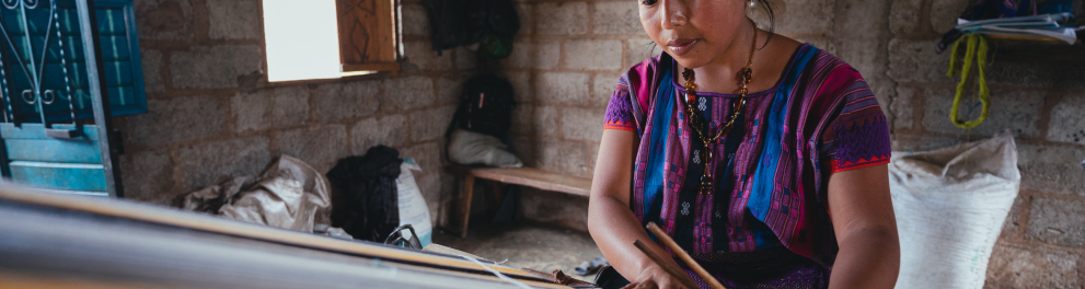 Mulher indígena com cabelos pretos e presos está detro de uma casa fazendo algum trabalho artesanal com peças típicas de sua cultura