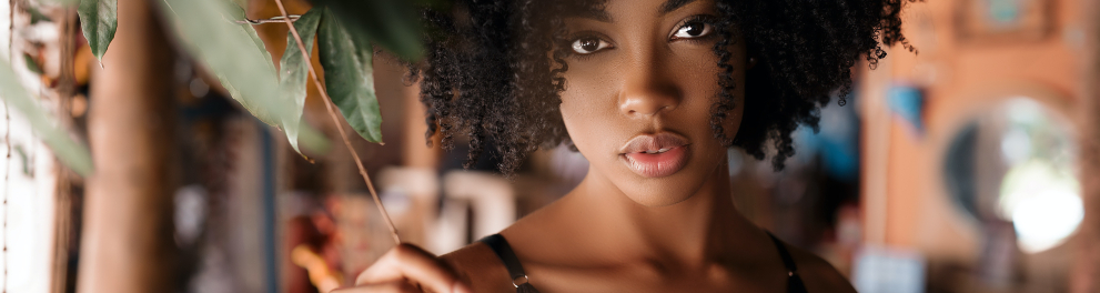 Uma linda e jovem mulher negra de cabelos cacheados no estilo black power. Ela está bem maquiada, olhando diretamente para a câmera e tocando uma plantinha com a mão direita