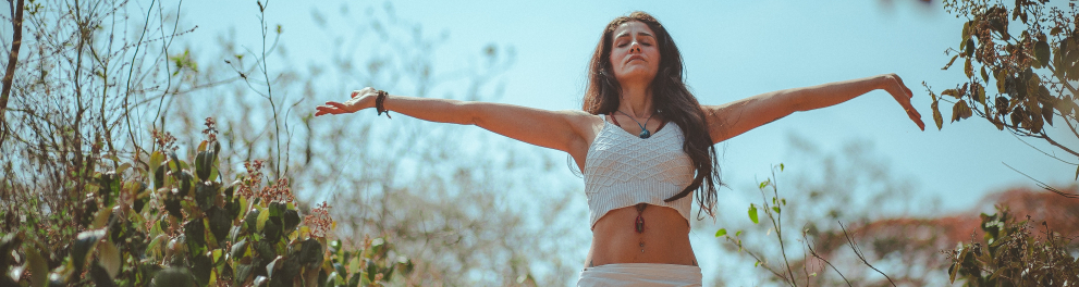 Uma jovem mulher branca está em um campo aberto com árvores, galhos e folhas ao seu redor, com o céu azul ao fundo. A mulher está de olhos fechados e braços abertos, demonstrando muita paz e tranquilidade