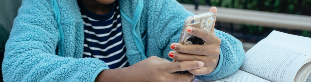 Imagem de uma menina negra bem sorridente, que está com um casaco azul, sentada e segurando e olhando para um celular com as duas mãos.