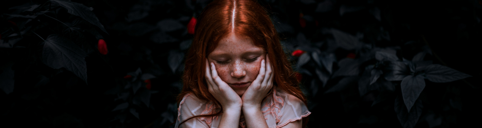 Uma menininha ruiva e sardenta, de cabelos batendo no ombro, está no centro da imagem com as duas mãos nas bochechas e olhando pra baixo, demonstrando tristeza. Ao fundo, a imagem está escurecida, mas nota-se que é um jardim 