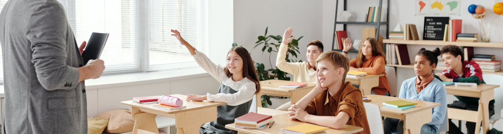 Imagem de uma sala de aula com algumas crianças sentadas em suas cadeiras e o professor em pé de frente para elas e de perfil para quem bateu a foto
