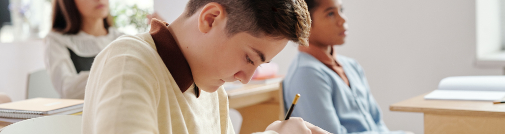 Menino branco trajando um suéter de cor creme está sentado em sala de aula e escrevendo algo em seu caderno com a mão direita. Ao fundo, podemos notar outras duas estudantes, colegas de classe do menino
