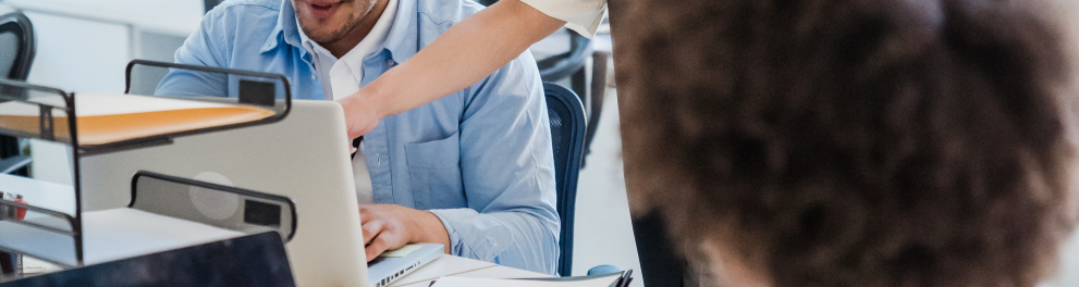 A imagem mostra uma mulher branca e loira em pé apontando para a tela do notebook de um rapaz branco que está sentado à mesa. Ela parece estar auxiliando ela com alguma dúvida de tecnologia da informação. à frente de ambos na imagem há outra mulher, negra de cabelos curtos e encaracolados, sentada e mexendo em seu próprio notebook de costas para quem bateu a foto