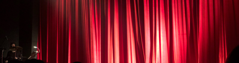 Imagem de uma cortina vermelha em um teatro. Abaixo do palco dá para ver algumas silhuetas de cabeças, o que mostra que há plateia sentada
