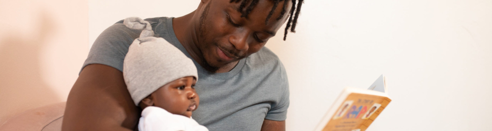 Um homem negro está com um bebê no colo e um livro em sua mão esquerda. Ele parece ser o pai da criança e está lendo para ela
